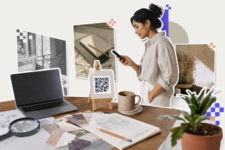 Designer at a desk holding a phone toward a printed card with a QR code on a small easel, a laptop and sketches beside it