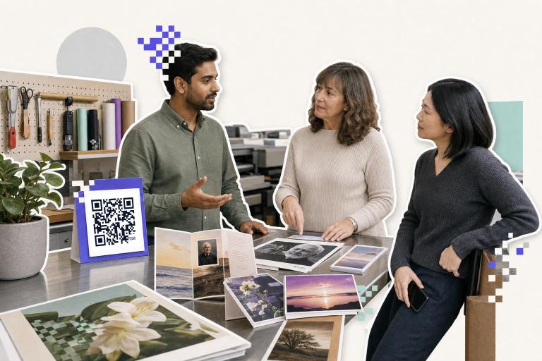 Three people discuss printed photos on a table with a QR code card nearby in a workshop setting.